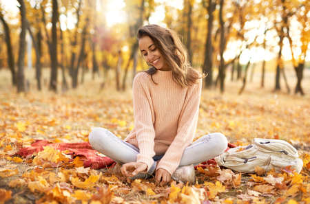 pretty woman in cozy sweater in autumn park with fall colorful background, sitting on blanket. Fall season concept. Outdoor lifestyle portrait.の写真素材