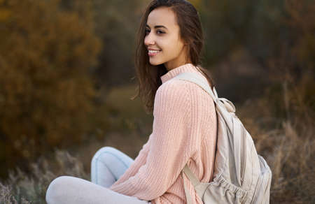 Closeup portrait woman in warm cozy sweater with backpack sitting and admiring nature in autumn forest. Fall season concept.の写真素材