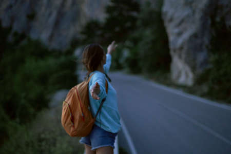 back view girl with backpack stands on road with mountains on background at evening, Follow me, first person view, focus on handの写真素材