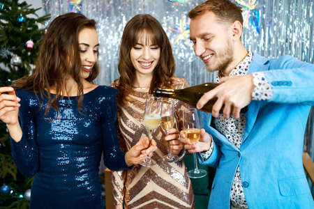 happy festive friends celebrating christmas or new year together and looking happy, pouring sparkling vine into glassesの写真素材