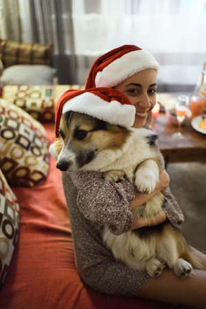 adorable happy woman and welsh corgi dog in in Christmas Santa hats hugging on sofa at home at Christmas timeの写真素材