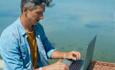 Concentrated man working outdoors by blue sea on laptop computer, searching information in social networks, keyboarding text. Concept remote work, freelanceの写真素材