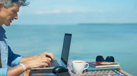 Panoramic image businessman working outdoors by beautiful blue sea view , searching information on laptop computer, keyboarding text. Concept remote work and travelの写真素材
