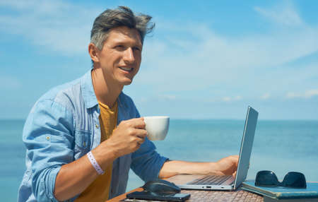 Portrait optimistic smiling man freelancer working outdoors on beach by blue sea, drinking coffee, using laptop computer. Dream office job workplace.の写真素材