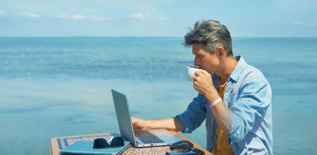 Pensive man freelancer working on laptop computer at beach by blue sea, drinking coffee. Concept remote work, freelanceの写真素材