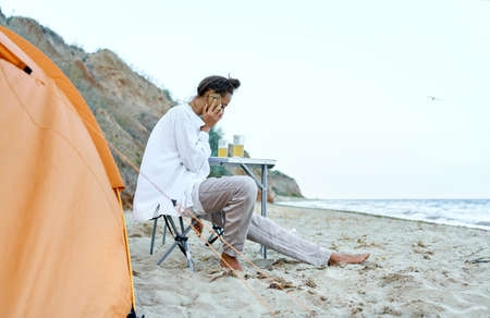 Traveler woman sitting by orange tent and cell phone calling, enjoying camping life at wild beach. Concept communication outdoorsの写真素材