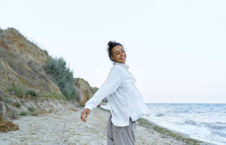 Playful carefree girl in white blowing shirt dancing at wild beach with eyes closed. Concept freedom and authentic emotionの写真素材