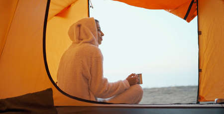 Reaer view carefree woman in funny pajama like bunny sitting at tent entry and holding mug with coffee in hand, enjoys morning in camp during travelの写真素材