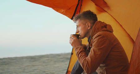 side view of adult man traveler sitting at tent entry, holding nd smelling mug with coffee in hands. man taveler drinking coffee at morning in camp during travel and enjoying calm landscapeの写真素材