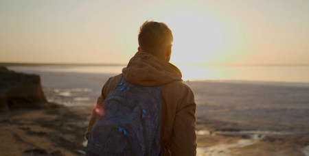 Back view man traveler with backpack walking along salt lake coast and meeting sunrise at deserted sea beach, sightseeing and looking around, enjoying freedom, fresh air and nature beautyの写真素材