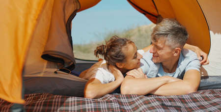Happy joyful travelers man and woman lying and relaxing in orange tent. Cute moments couple in love during vacation or weekend, enjoys free camping lifeの写真素材