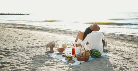 Rear view couple having picnic at the beach, sitting on blanket and embracing, looking to seaviewの写真素材