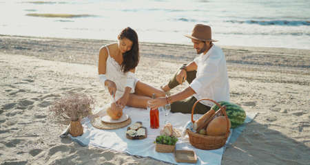 Beautiful couple having romantic breakfast with lots of tasty food and wine, sitting on piknic planket at beach with ocean viewの写真素材