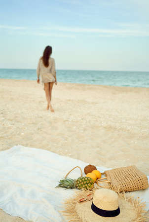Summer fashion beach accessories and fresh fruit on sandy sea beach, silhouette walking woman on background. summer time and holiday outdoors concept.の写真素材
