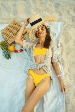 joyful happy woman in yellow bikini lying on blanket at beach next to beach accessories and fruitの写真素材