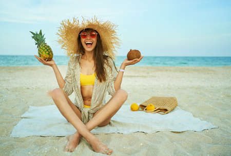 Laughing happy woman in red eyeglasses wearing straw hat, holding tropical fruit on beachの写真素材