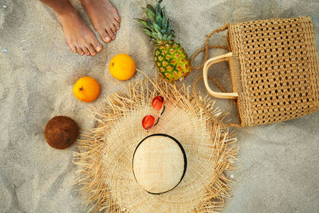 Top view summer female feet next to fashion outfit and tropical fruit flat lay on sand