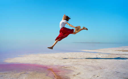 Happy woman in red skirt jumping, enjoying bright pink salt lake with blue skyの写真素材