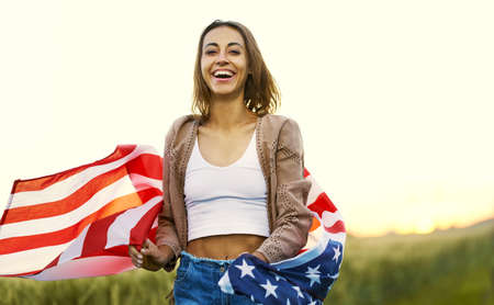 Laughing girl enjoying nature at sunset in summer, running at field with American flagの写真素材