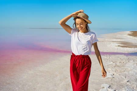 Joyful tourist woman in red skirt and hat walking by salt flats beach at pink lakeの写真素材