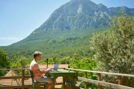 Side view of man freelancer working on laptop computer on terrace with beautiful mountain nature view. Technology and travel, freelance conceptの写真素材