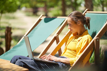 Portrait skilled freelance woman using laptop computer while sitting on sun lounger outdoors, searching information, keyboarding text, online working digitalの写真素材