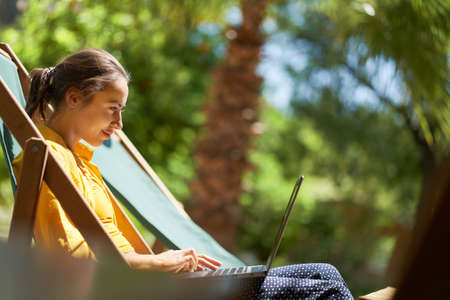 Side view remote online working woman sitting on sun lounger with palm treesの写真素材