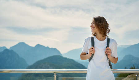 Pleased tourist female wearing white shirt with backpack standing in front of mountain top with cloudy sky, viewpoint of Tunektepe Teleferik, Antalyaの写真素材