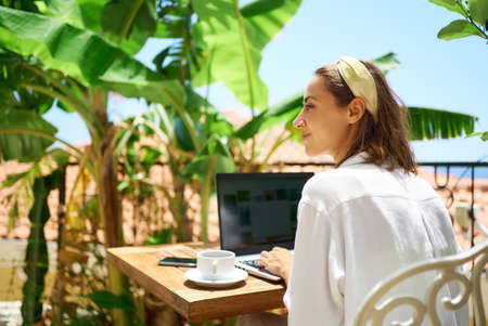 Young beautiful mixed race woman sitting on terrace at morning in hotel, using laptop computer while enjoying vacationの写真素材