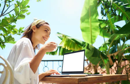 Cheerful freelancer womam drinking cofee at morning at balcony with palm trees, sitting with laptop blank screen. Work and vacationの写真素材