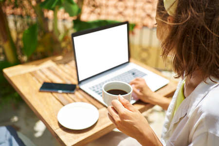 Rear view crop faceless female working on laptop computer with white empty screen. typing, browsing, searching information while drinking coffeeの写真素材