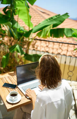 Rear view female working remotely on the laptop computer, typing keyboarding text while sitting on balcony hotel with palm treesの写真素材
