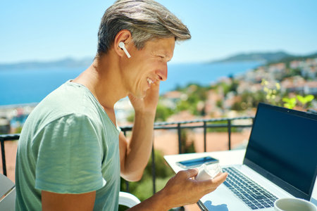 Close-up portrait happy smiling man wearing headphones, looking at laptop while sitting at terrace...