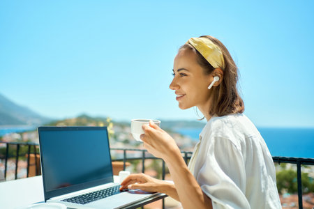 Young business woman drinking coffee and looking away, working on laptop computer at morning while sitting at terrace with sea view on resortの写真素材