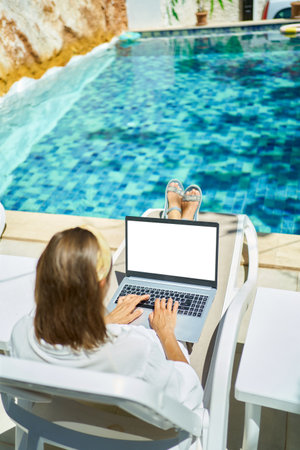 Rear view female working remotely on the laptop computer, typing keyboarding text while lying on sun loungers by swimming poolの写真素材