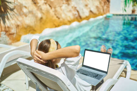 back view woman relaxing chilling by the pool during vacation at resort hotel, watching news or video on laptop empty screenの写真素材