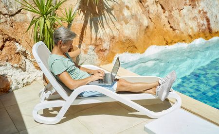 Adult man relaxing on lounger by the swimming pool during vacation at resort, typing on keyboard,...