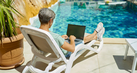 Rear view man relaxing on lounge by the swimming pool at resort hotel, using laptop computer, typing text on keyboard, work and travel conceptの写真素材