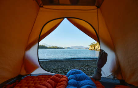 Camping with tent at beach of Mediterranean sea, two tourists people lying in sleeping bag with view on seashoreの写真素材