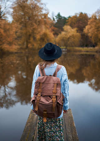 Rear view traveler woman in hat with backpack standing on the promenade at lake, looking at autumn nature view, trees with bright orange foliage reflection in waterの写真素材