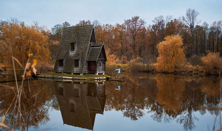 Panoramic image autumn scenic landscape. Wooden house by lake among forest, girl walking by wooden pier. foliage reflection in water.の写真素材