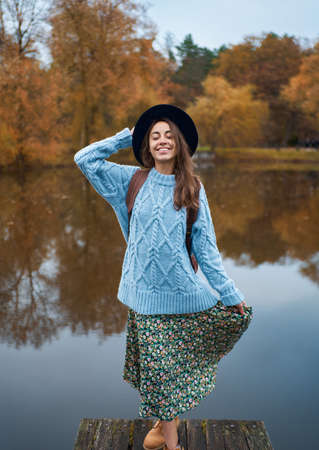 Joyful traveler woman in warm blue sweater and hat standing on the pier at lake with autumn nature view, trees with bright orange foliage reflection in waterの写真素材