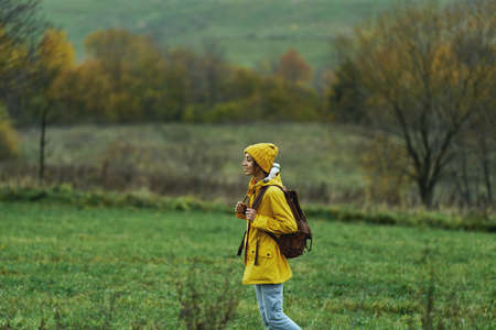 Woman hiker with backpack walking by green meadow among hills, girl wearing yellow jacket and hat. Travelling in fall seasonの写真素材