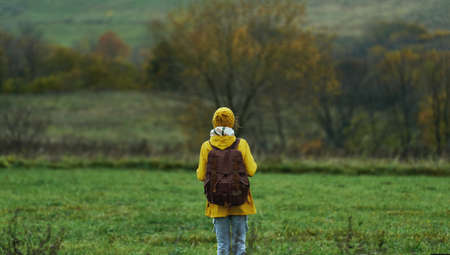 Back view female tourist with backpack standing alone at green lawn among hills. Cold weather, misty autumn nature viewの写真素材