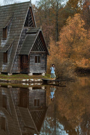 Old wooden house hut by lake among autumn forest, woman walking by wooden pier. Foliage reflection in water.の写真素材