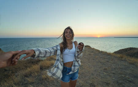 Happy positive emotions girl laughing and holding hand of her friend on cliff with seascape during sunset. Adventure lifestyle, love travelingの写真素材