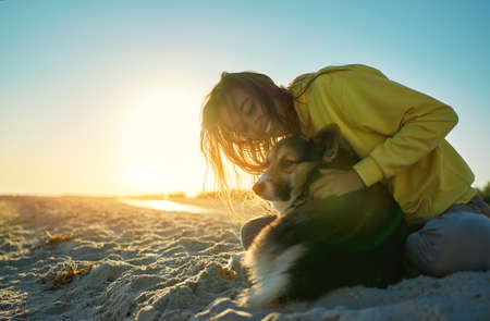 Happy woman in morning sun light with her pet at sandy beach. Girl hugging cute Welsh Corgi dogの写真素材