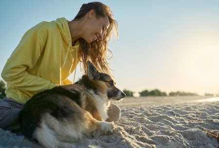Joyful smiling woman with her pet Welsh Corgi dog spending time together at sandy beach with morning sunshineの写真素材
