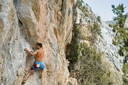 Strong man with muscular body climbing on vertical cliff with beautiful lines and stone texture, climber hiker working out outdoors with mountains viewの写真素材