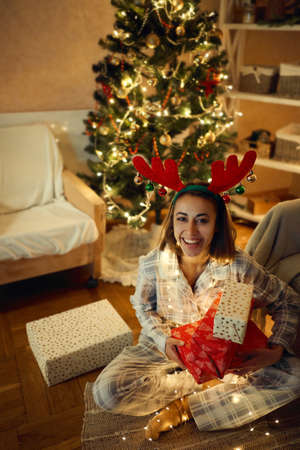 Cute cheerful woman sitting on floor at home by Christmas tree, embracing gift boxes. Happy girl wearing reindeer antlers headband and cozy pajamasの写真素材